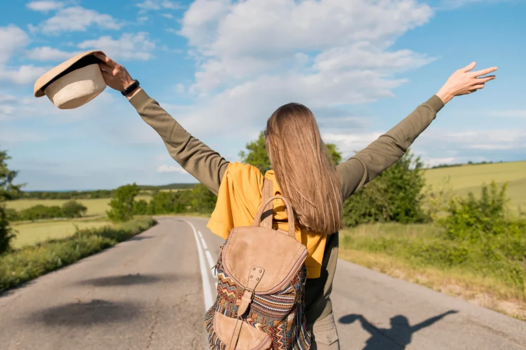 Caminos de autodescubrimiento para aumentar la confianza 1 Mujer con los brazos levantados y un sombrero en la espalda, de pie junto a la carretera, expresando alegría y libertad.