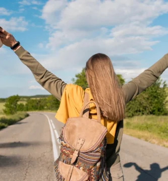 Mujer con los brazos levantados y un sombrero en la espalda, de pie junto a la carretera, expresando alegría y libertad.