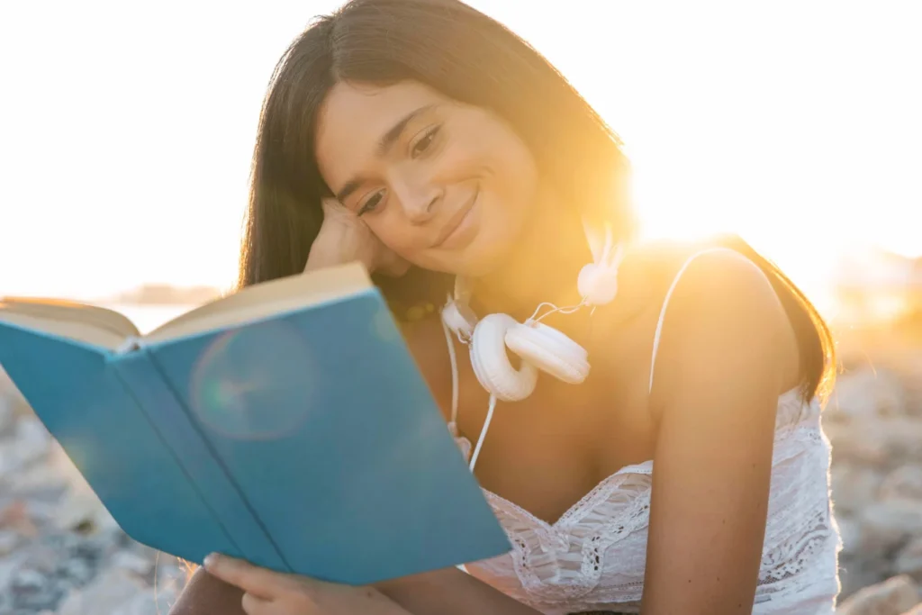 Mujer en la playa, usando auriculares y leyendo un libro, sumergida en un momento de tranquilidad junto al mar.