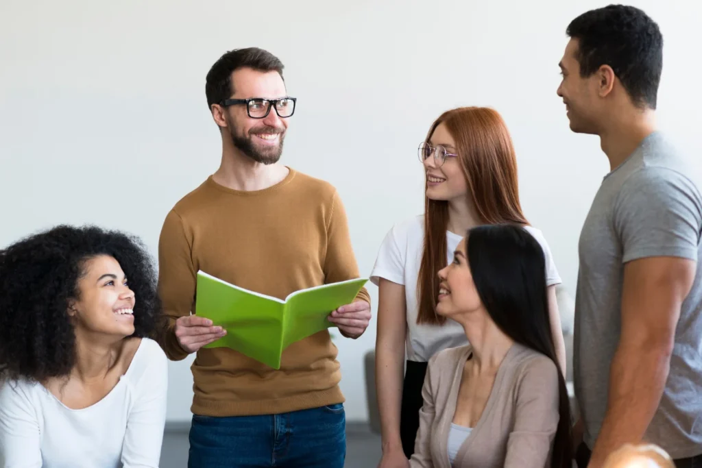 10 Consejos para Iniciar una Conversación con Confianza 3 Un hombre que sostiene una carpeta verde abierta sonríe y habla con cuatro personas que escuchan atentamente y sonríen a su alrededor en un ambiente informal.